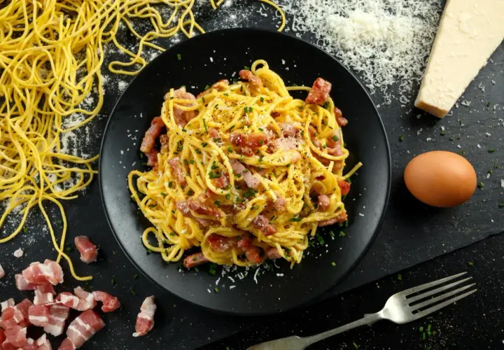 Pyszne spaghetti carbonara na talerzu, obok surowy boczek, jajko i ser. Zastanawiasz się, ile kalorii ma spaghetti carbonara?