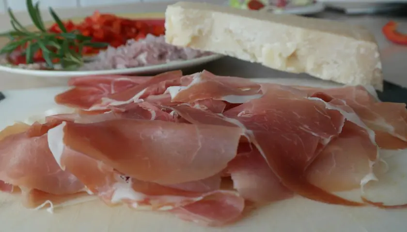 Cienko pokrojona szynka crudo, kawałek parmezanu i talerz z dodatkami.