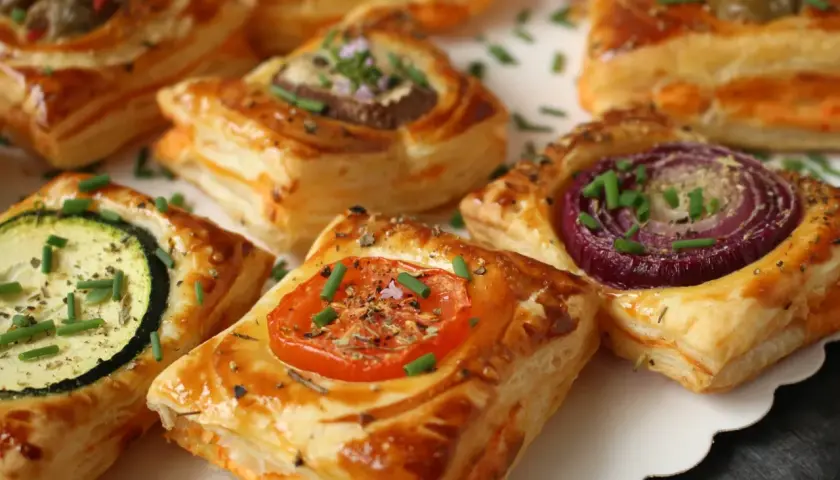 Kwadratowe ciastka francuskie z warzywami: pomidorem, cukinią, cebulą i oliwkami, posypane szczypiorkiem.