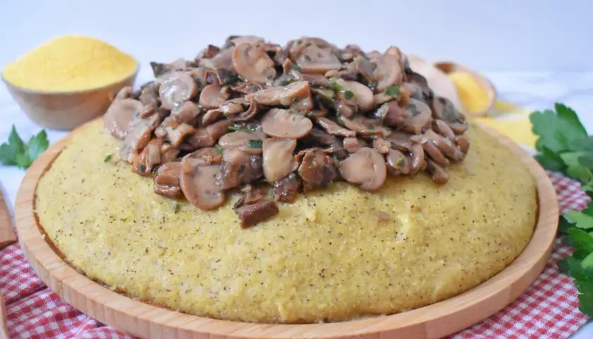Polenta z duszonymi pieczarkami na drewnianej desce, obok miseczka mąki kukurydzianej.