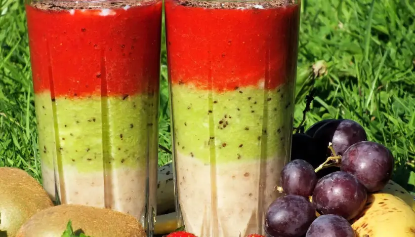 Dwa warstwowe koktajle owocowe w szklankach, otoczone owocami: kiwi, truskawki, winogrona i banan.