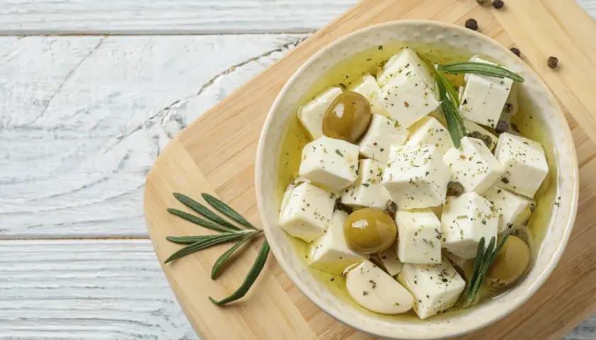 Kostki sera feta w oliwie z oliwkami, czosnkiem i rozmarynem na desce.