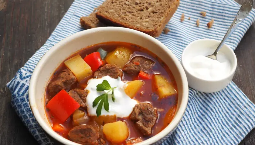 Gęsty, aromatyczny **бограч** z kawałkami wołowiny, ziemniaków i papryki, udekorowany śmietaną i natką pietruszki. Obok kromka chleba i miseczka śmietany.