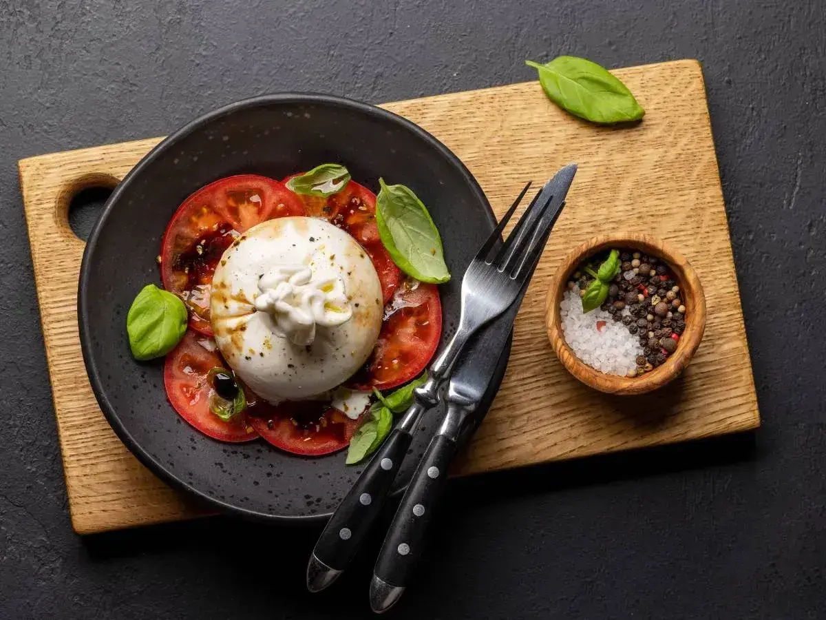 Pyszna burrata z pomidorami, polana oliwą, ozdobiona bazylią. Obok miseczka z solą i pieprzem.