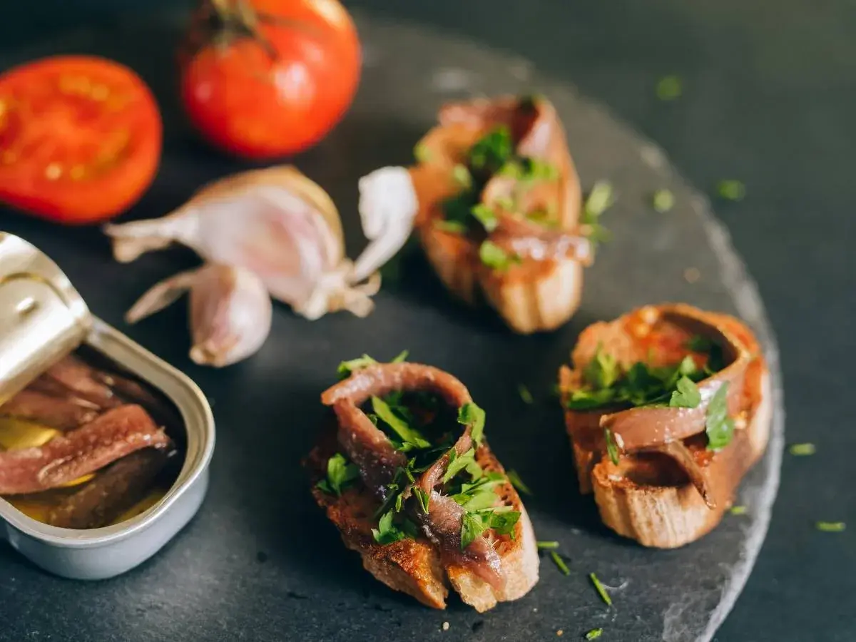 Otwarta puszka anchois, bruschetty z anchois i natką pietruszki, pomidory i czosnek na ciemnym tle.