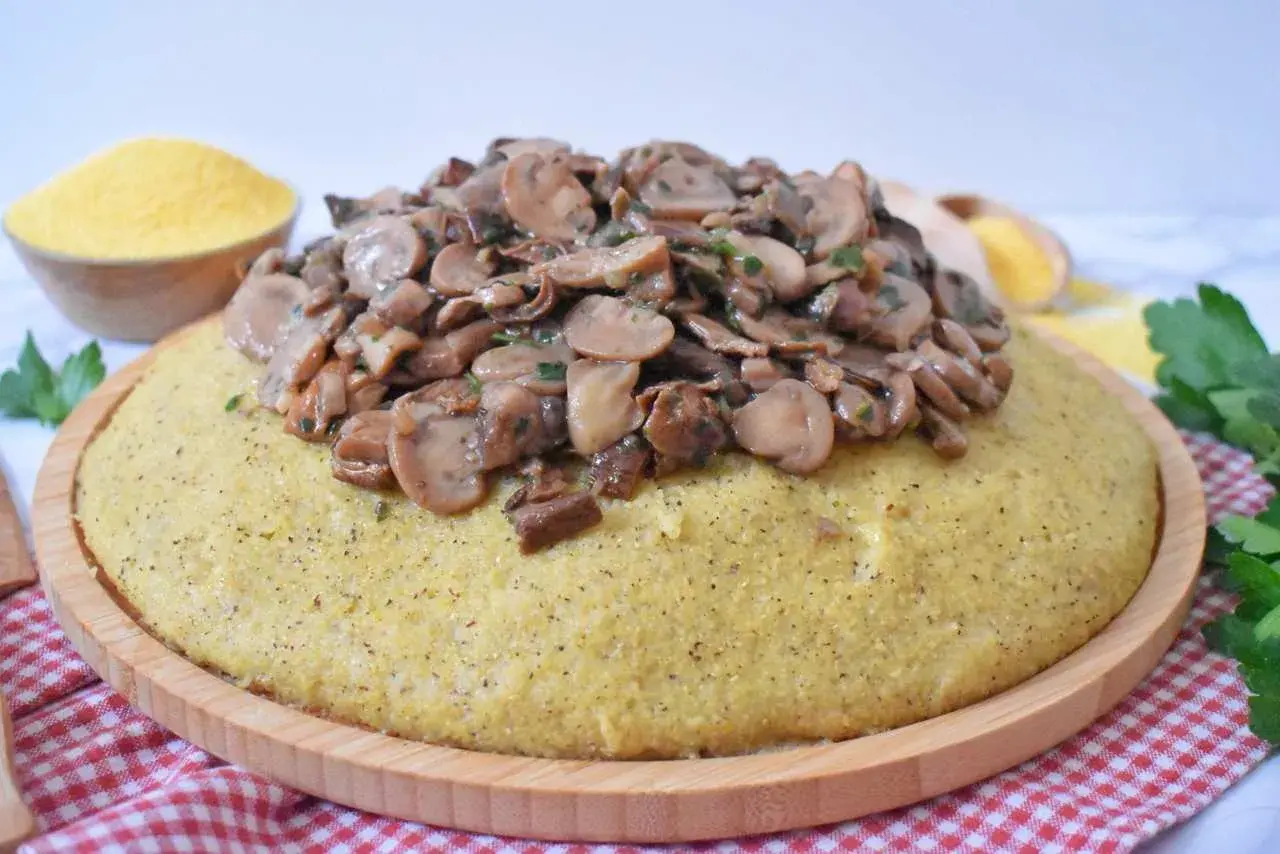 Polenta z duszonymi pieczarkami na drewnianej desce, obok miseczka mąki kukurydzianej.