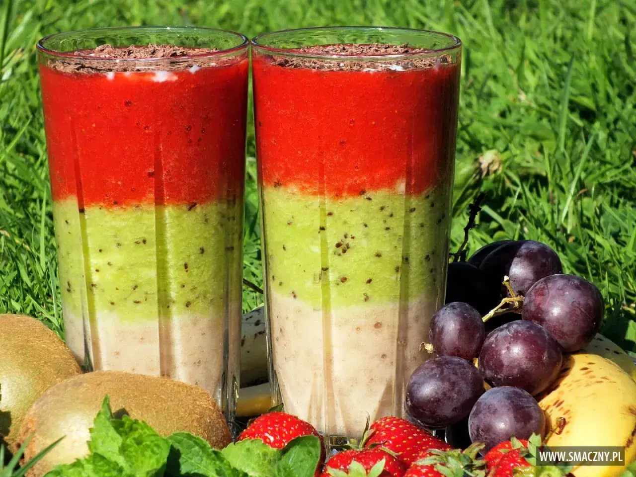Dwa warstwowe koktajle owocowe w szklankach, otoczone owocami: kiwi, truskawki, winogrona i banan.