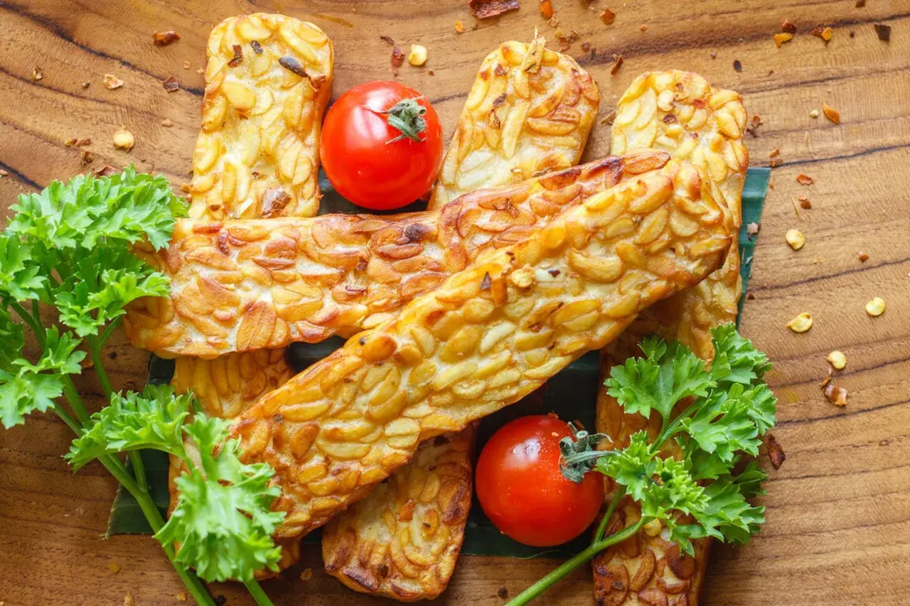 Usmażony tempeh z pomidorkami koktajlowymi i natką pietruszki na drewnianym tle.