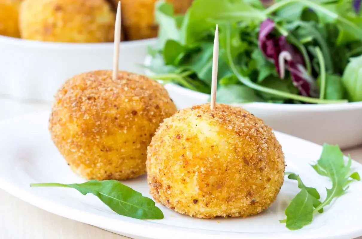 Dwa złociste arancini z wykałaczkami na białym talerzu, obok sałatka.