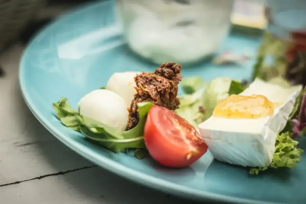 Keto śniadanie: ser brie z dżemem, kulki mozzarelli, pomidor i rukola na niebieskim talerzu.