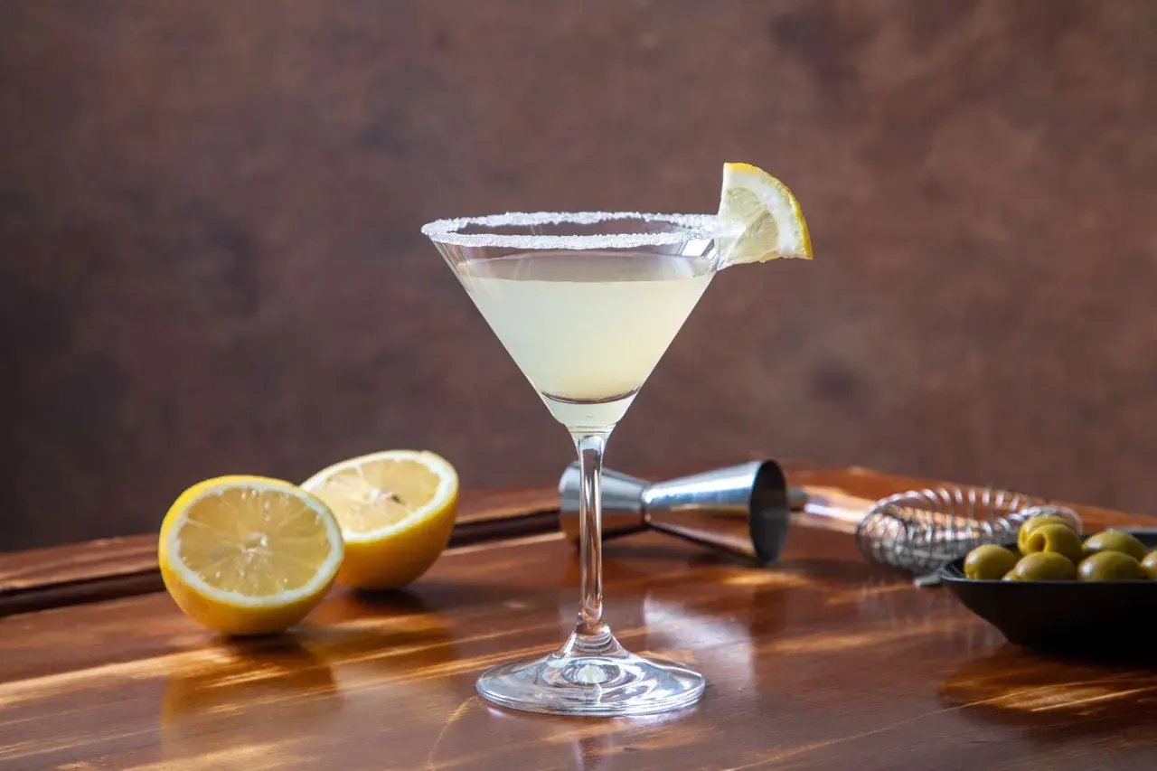 Elegancki martini bianco drink z solnym rantem, ozdobiony plasterkiem cytryny. Obok leżą połówki cytryn i miseczka oliwek.