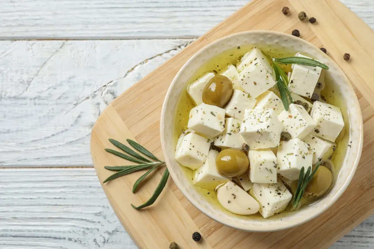 Kostki sera feta w oliwie z oliwkami, czosnkiem i rozmarynem na desce.