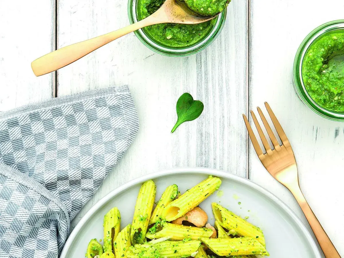 Makaron penne z zielonym pesto, podany na talerzu, obok słoików z pesto i złotego widelca.