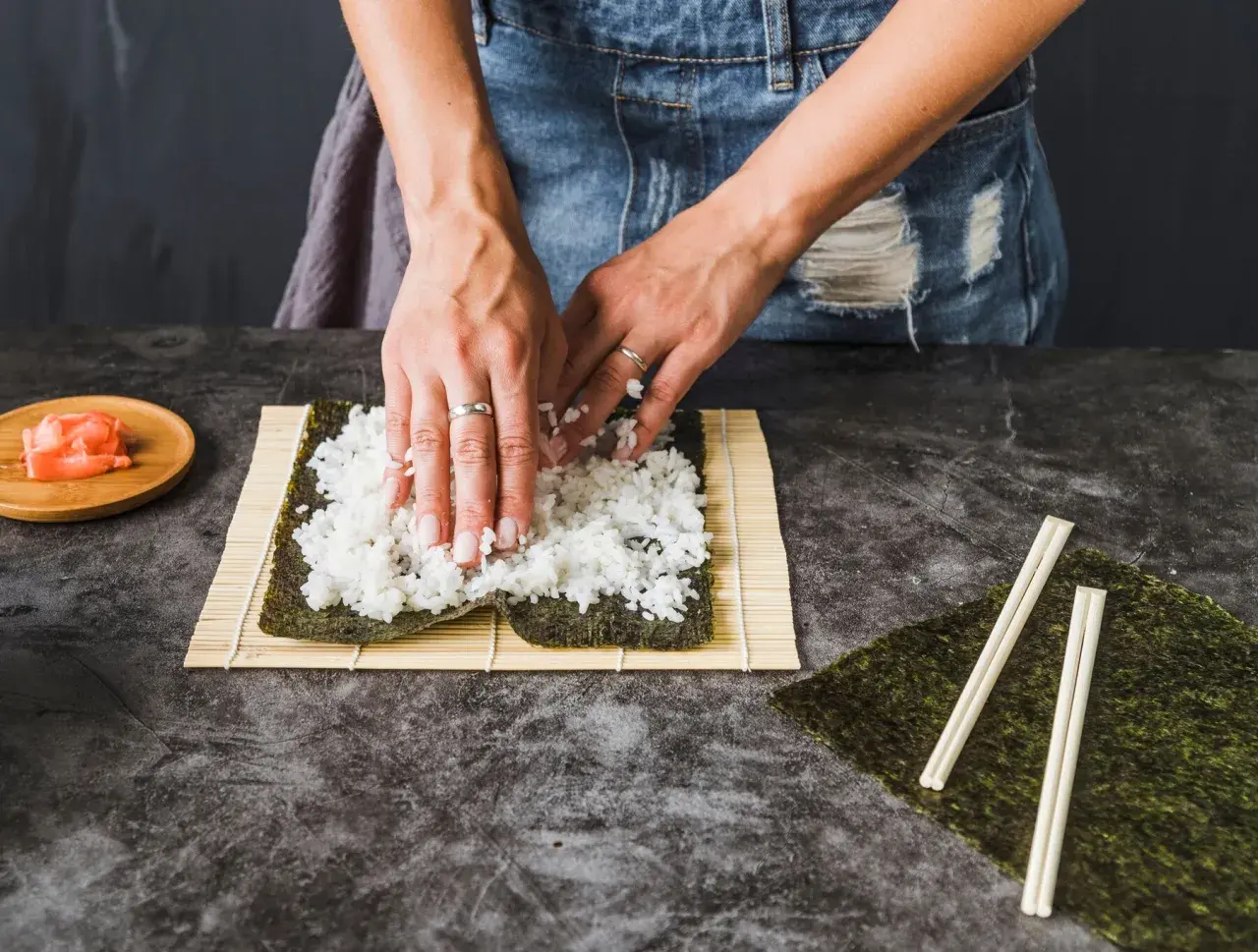 Dłonie rozkładają ryż na nori na macie bambusowej do sushi.
