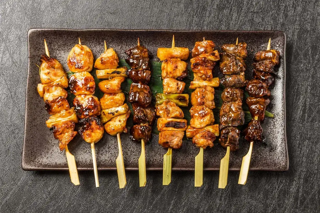 Różnorodne yakitori na patyczkach, z kurczakiem i warzywami, błyszczące od sosu, ułożone na ciemnej tacy.