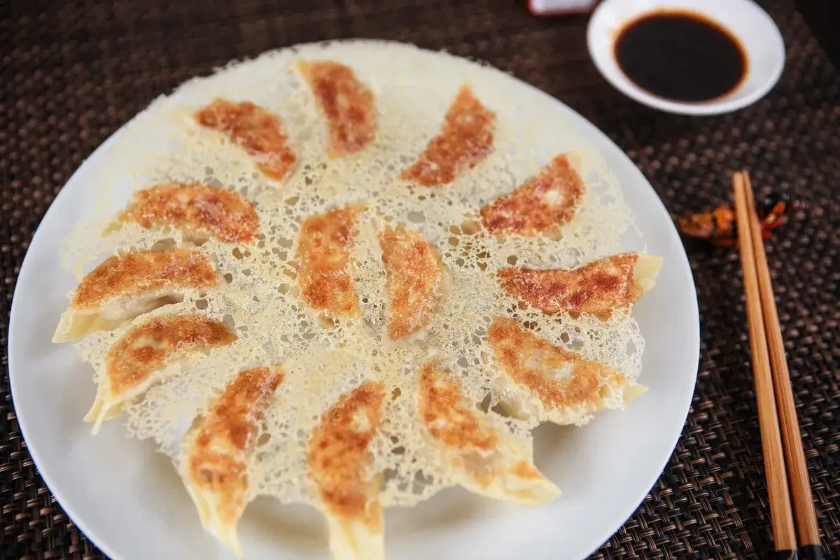 Chrupiące pierożki gyoza ułożone na talerzu, otoczone delikatną, złotą panierką z sera. Obok miseczka sosu sojowego i pałeczki.