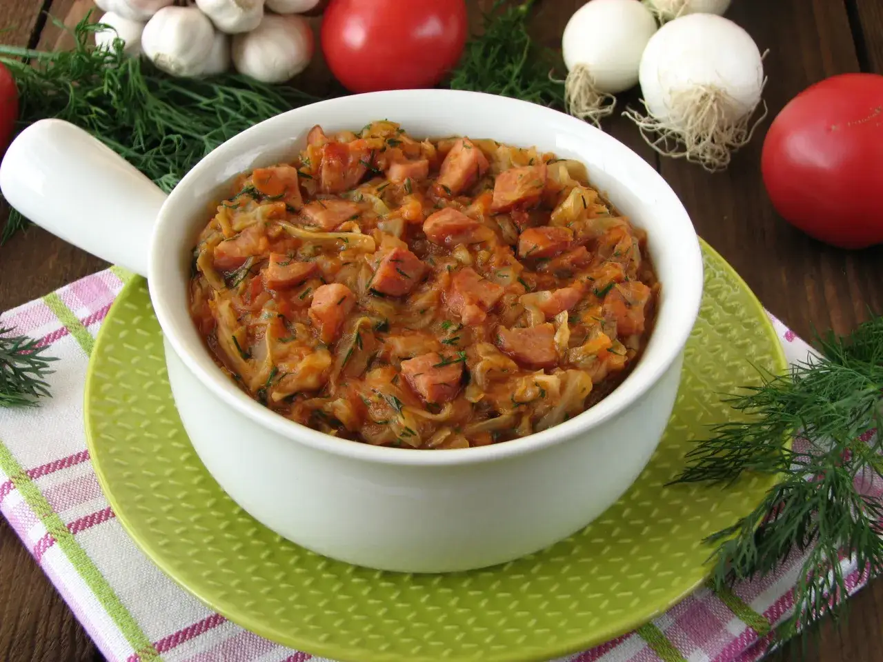 Jak ugotować dobry bigos z młodej kapusty? Aromatyczne danie z kapusty i kiełbasy w białej misce, na zielonym talerzyku.